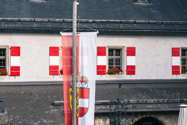 The red and white Salzburg banner with its crowned coat of arms hangs from a timber mast in the courtyard of Festung Hohenwerfen, in front of a whitewashed range with red-striped shutters and geraniums.