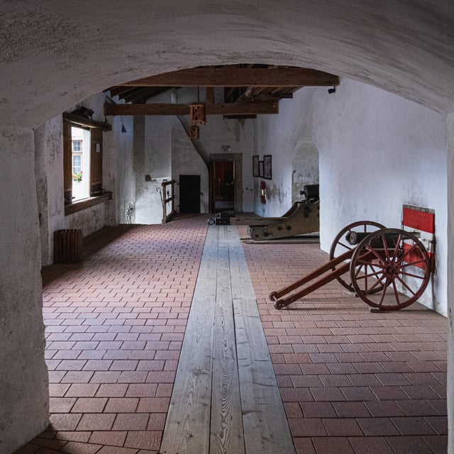 A long barrel-vaulted passage at Festung Hohenwerfen with tiled floor and a central plank run, a red-wheeled field cannon parked against the right wall and further gun carriages beyond.