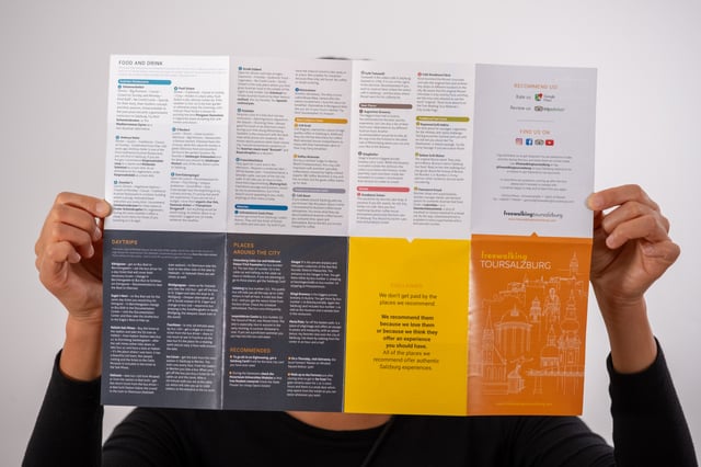 Two hands hold the Free Walking Tour Salzburg city guide open at full spread, showing the food and drink recommendations, the day trips column and the orange cover panel.