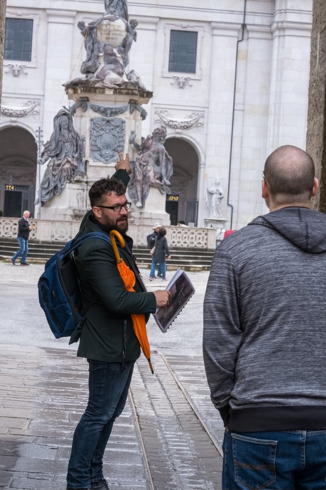 A tour guide with an orange umbrella turns and points up at the Marian column on Domplatz while a guest listens, the cathedral facade with its 774 and 1959 date panels behind.