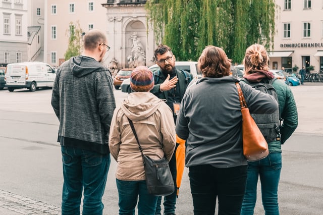 A tour guide speaks with an open hand to four visitors gathered on Kapitelplatz in front of the Kapitelschwemme horse pond and the Dombuchhandlung shopfront.
