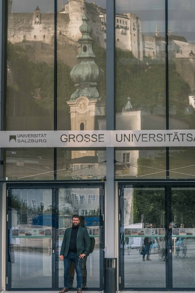Man in blazer stands at glass entrance of the Grosse Universitaetsaula with fortress reflected in the facade in Salzburg