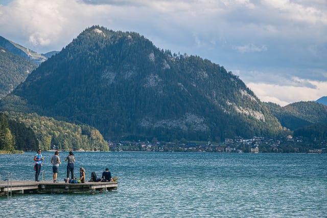 Five people stand and sit at the end of a low wooden jetty on Fuschlsee, with the village of Fuschl am See spread along the far shore beneath a steep forested peak.