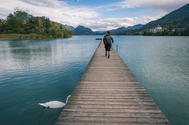A wooden jetty runs out into Fuschlsee with a swan feeding at its edge and a walker on the planks, while the yellow Schloss Fuschl resort stands among trees on the far right shore.