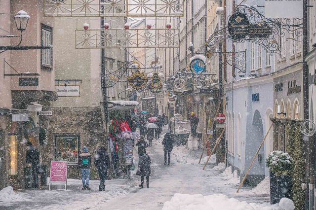 Snow falling on Getreidegasse with wrought iron guild signs and Christmas decorations in Salzburg old town.