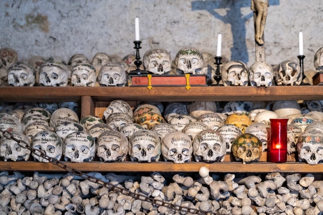 A wide view of the charnel house wall showing three tiers of painted skulls beneath a wooden crucifix and the blue cross painted on the whitewash behind it, with the lowest register filled by packed bones.