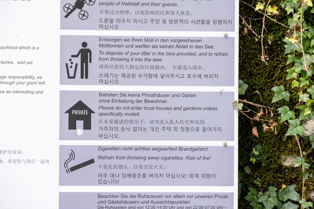 A close view of the visitor conduct panel on Hallstatt's information board, showing pictograms and multilingual text asking people not to fly drones, not to throw litter in the lake, not to enter private houses and gardens, and not to drop cigarettes.