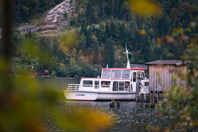 The small white passenger boat Stefanie lies at a wooden jetty on the Hallstaetter See, framed by out of focus autumn leaves, with a steep forested slope and rock face behind.