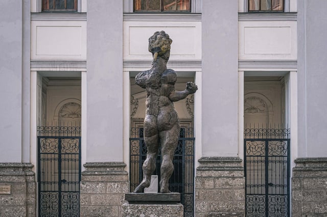 Homage to Mozart sculpture framed by the columned facade and iron gates of Markuskirche in Salzburg.