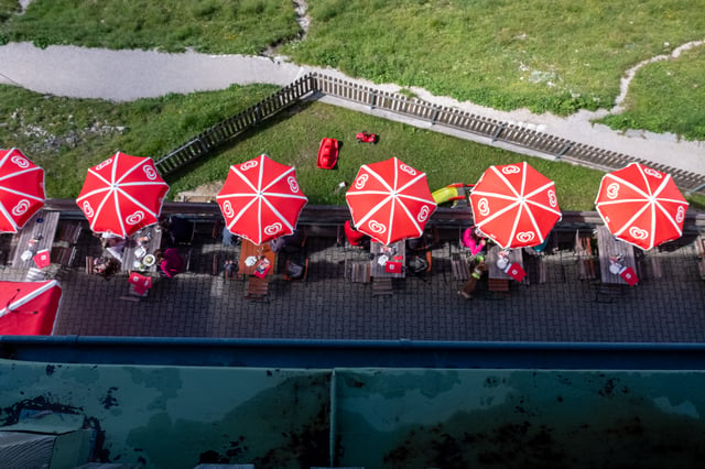 Looking straight down from an upper window onto a row of red and white parasols shading wooden benches on the paved summit terrace, with a picket fence and green slope beyond.