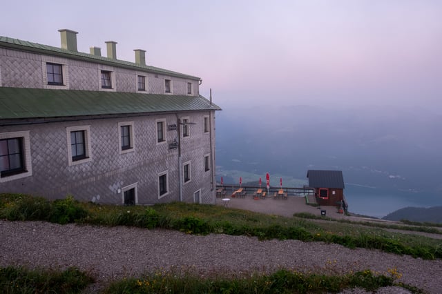 The long shingled flank and green metal roof of the Hotel Schafbergspitze at dawn, with its terrace of furled red umbrellas and a small kiosk hut looking down to the Wolfgangsee.