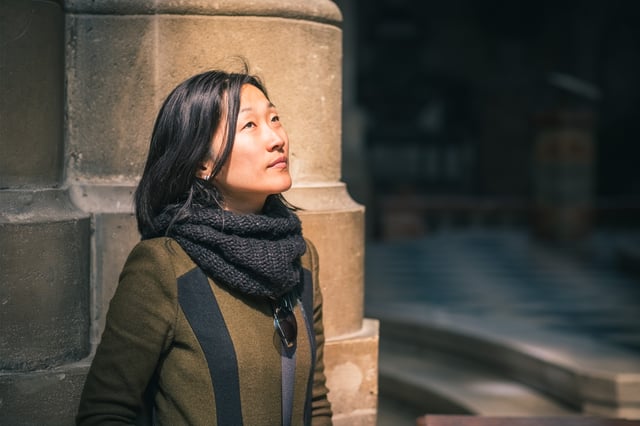 A woman in an olive coat and a black knitted snood stands against a sandstone cathedral pier in a shaft of sunlight, chin raised, looking up into the building.