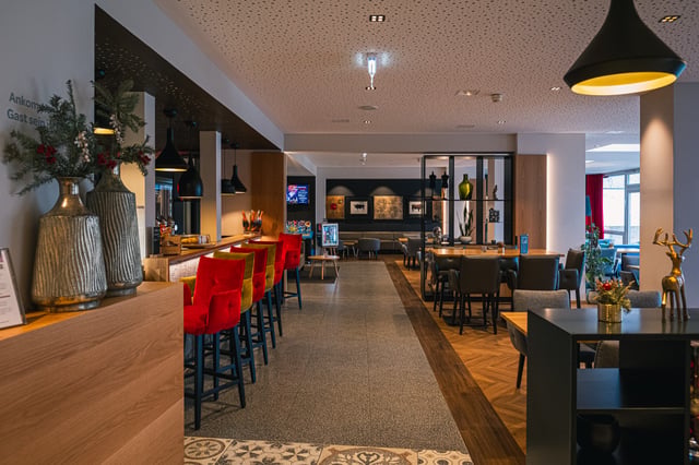 JUFA Hotel Salzburg, bar at Christmas, red and ochre velvet stools along a pale oak counter, a terrazzo aisle inlaid with a patterned tile panel and a dining room of dark chairs beyond.