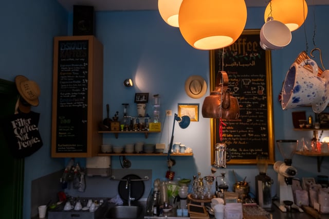 The bar of Kaffee Alchemie in Salzburg under orange globe lamps, with a copper kettle and a glass press hung on wires in front of the gold-framed coffee menu and an Eiskalte Kaffees chalkboard on the blue wall.