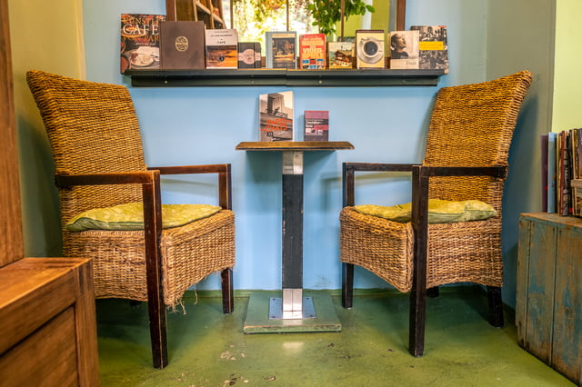 Two high-backed wicker armchairs with green cushions face each other across a small dark table in a corner of a coffee shop, below a shelf of books about coffee and cafés, against a pale blue wall on a green painted floor.