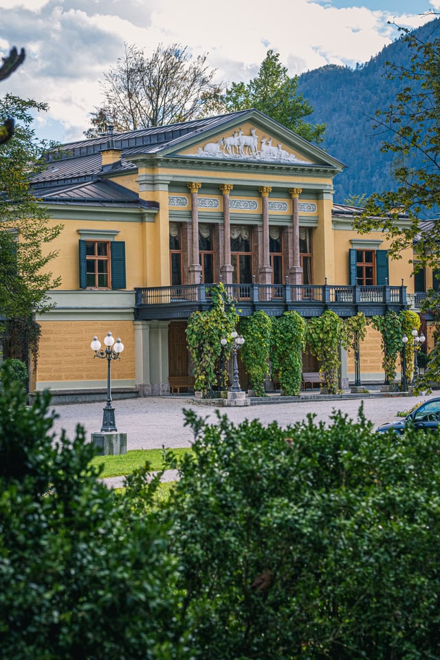 Upright view of the Kaiservilla's central portico, its four columns under a pediment filled with a white relief of deer, with a wooded mountain rising behind.