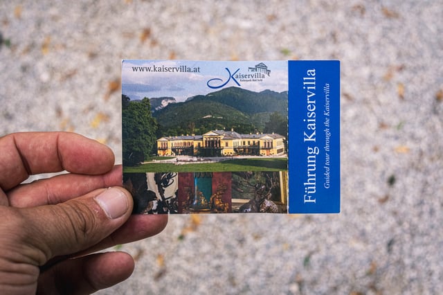 A hand holds a printed Kaiservilla guided-tour ticket showing the yellow imperial villa against wooded hills, above a gravel path in Bad Ischl.