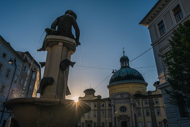 Fountain statue silhouette with Kajetanerkirche dome at sunset on Kajetanerplatz in Salzburg.