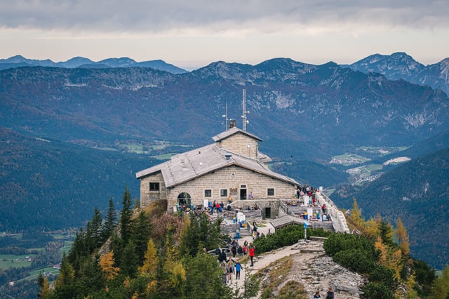 The Kehlsteinhaus sits on its rocky spur above the Berchtesgaden valley, its stone roof and aerial mast seen from the summit path with layered ranges behind.