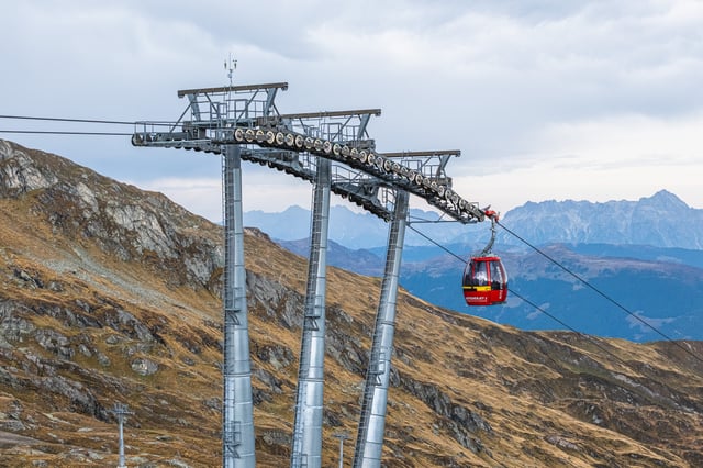 A red Gletscherjet 2 gondola of the Kitzsteinhorn lifts hangs between three tall steel pylons above brown autumn slopes, with limestone ranges fading into haze behind.