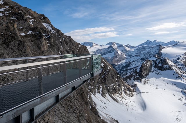 The steel skywalk gangway at the Kitzsteinhorn summit runs out along the cliff edge, with glaciated Hohe Tauern peaks and one sharp dark summit on the horizon beyond.