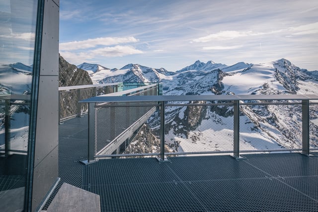 The empty grated terrace at the Kitzsteinhorn summit, its mesh railings framing a panorama of glaciated Hohe Tauern peaks under high thin cloud.