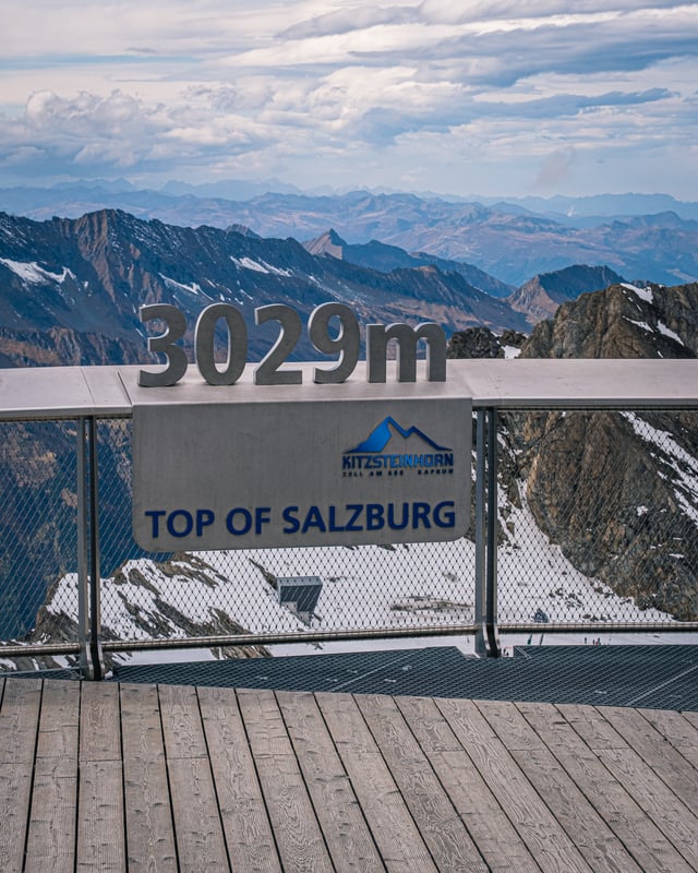 Steel numerals reading 3029 m stand on the railing of the Kitzsteinhorn summit platform above a panel lettered Top of Salzburg with the resort logo, snow-streaked peaks of the Hohe Tauern beyond.