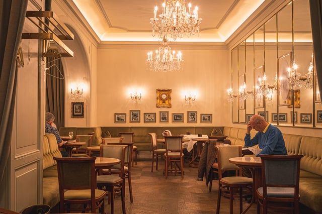 The warmly lit cafe salon at Zauner in Bad Ischl, with crystal chandeliers, olive banquettes, bentwood chairs and two guests reading at marble tables.