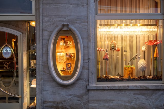 The lit shop window of Zauner in Bad Ischl at dusk, filled with a display of oversized sugar and marzipan toadstools beside a marble niche of pralines.