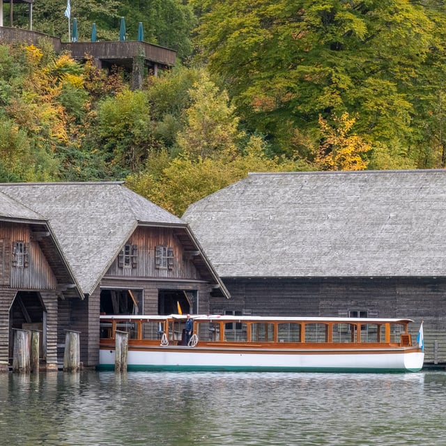 A long white and varnished electric passenger boat lies moored under the shingle-roofed timber boathouses at the Königssee landing, autumn beeches rising behind.