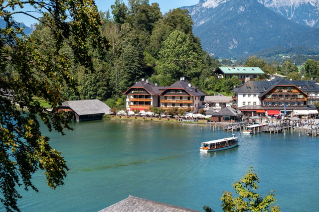 Framed by overhanging branches, an excursion boat pulls away from the Koenigssee jetties past a dark timber boathouse, the turquoise water tracing its wake.
