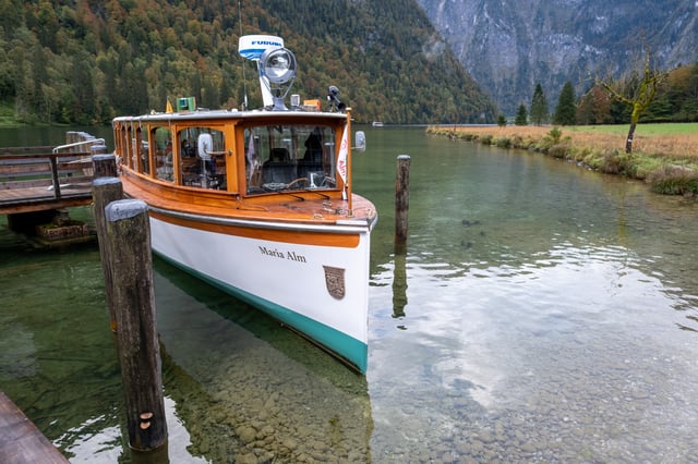 The electric tour boat Maria Alm lies bow on at the wooden jetty on the Koenigssee, its varnished hull above water so clear the stones on the bottom are visible.