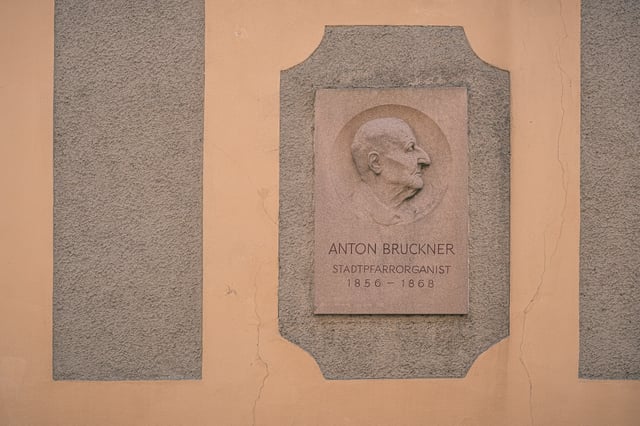 A stone memorial relief on an ochre wall shows Anton Bruckner in profile above the inscription Stadtpfarrorganist 1856 to 1868.