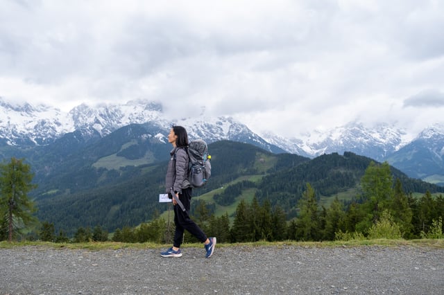 A hiker with a large grey backpack walks along a gravel track above Maria Alm, a long wall of snow-covered limestone peaks under heavy cloud filling the horizon behind her.