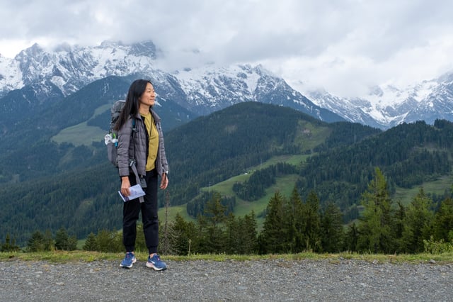 A hiker stands in profile holding a folded leaflet and looks out across the wooded Urslau valley toward the snow-covered range above Maria Alm.