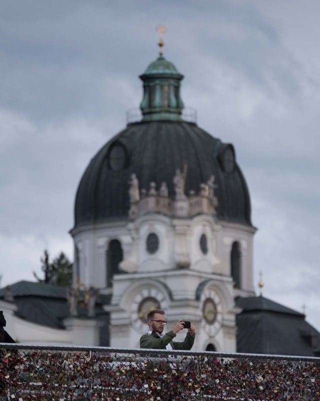 Man taking a photo on Marko Feingold footbridge with Collegiate Church dome behind in Salzburg.