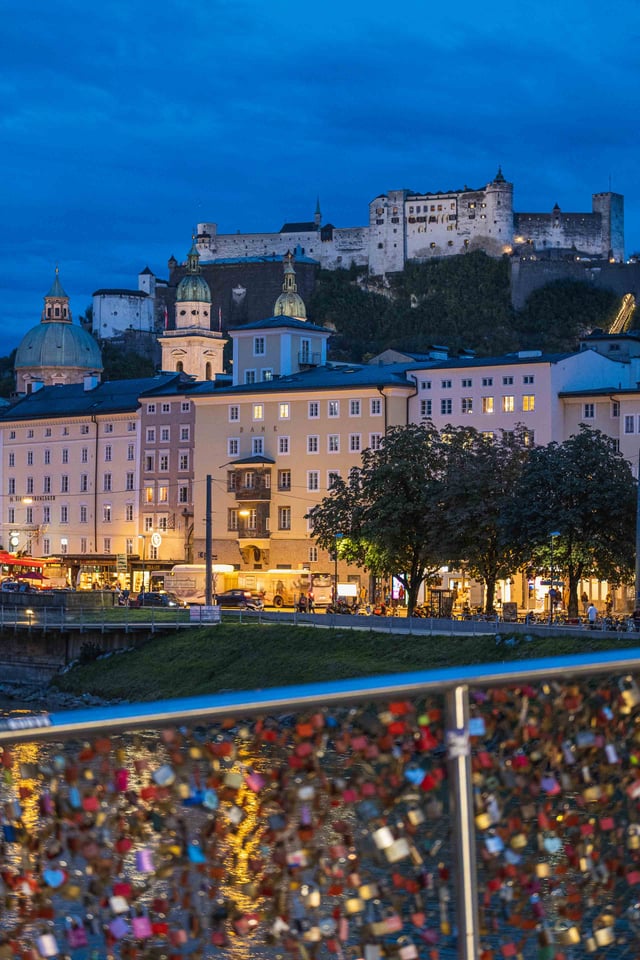 Love locks on Marko-Feingold-Steg railing with Hohensalzburg Fortress lit at dusk behind