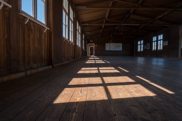 Sunlight from a row of windows falls in bright rectangles across the bare plank floor of an empty wooden prisoner barracks at the Mauthausen Memorial.