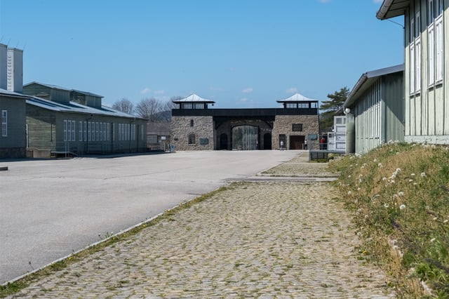 The stone gatehouse of the Mauthausen Memorial with its two watch platforms, seen across the empty roll call square between the flanking barracks.