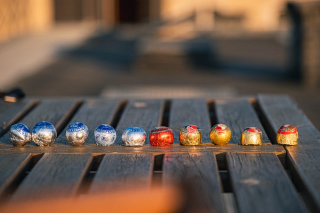 Ten Mozartkugeln from different makers lined up in a single row across the slats of a wooden deck table in low evening sun.