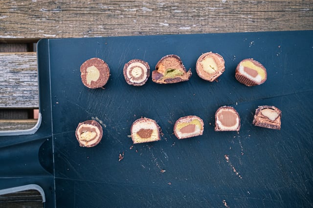 Ten Mozartkugeln cut in half and arranged in two rows of five on a dark cutting board, each cross section showing a different layering of marzipan, nougat and chocolate.