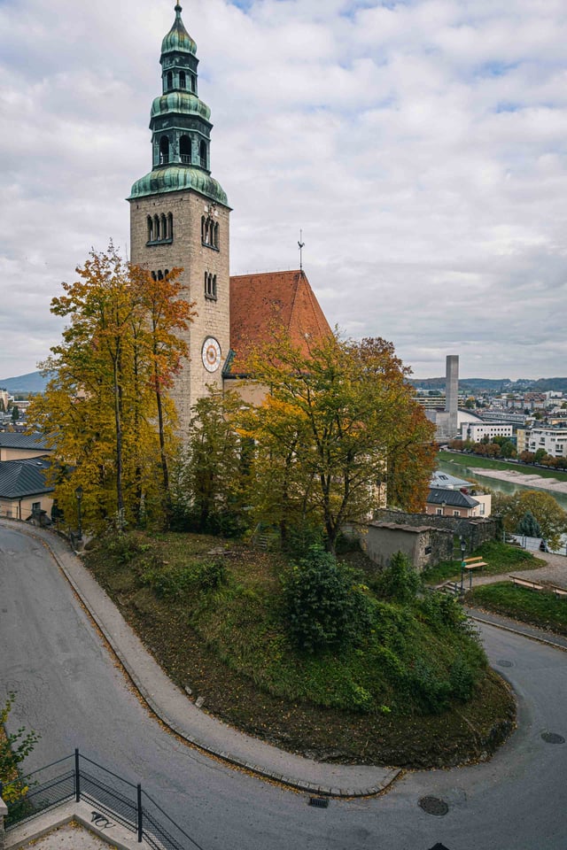 Muelln Church exterior with brick facade and green tower rises above autumn trees in Salzburg.