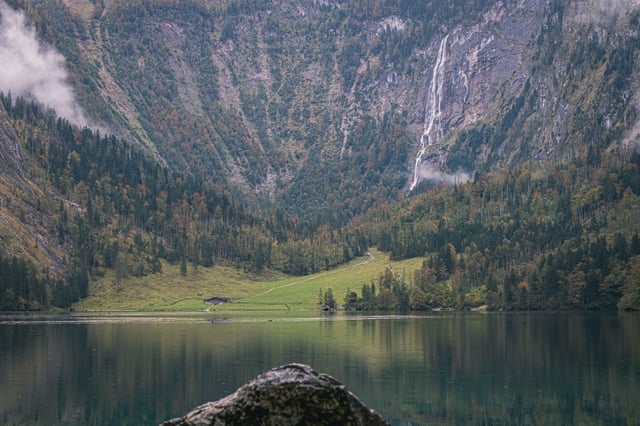 Seen from the Obersee shore over a lichen-covered boulder, the Rothbach waterfall falls beside the green Fischunkelalm meadow at the head of the lake.