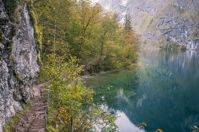 Looking down from the cliff path on the Obersee, the narrow track curves along the rock face beside water shading from clear shallows to deep turquoise.