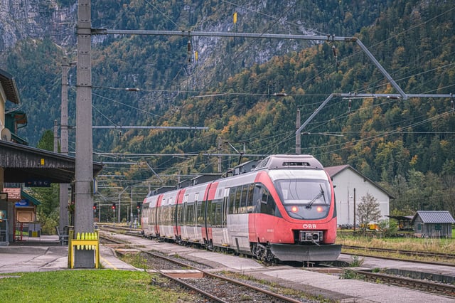 A red and white OeBB Talent railcar stands at the platform of the Obertraun station on the Salzkammergut line, with an autumn-coloured cliff face rising behind it.