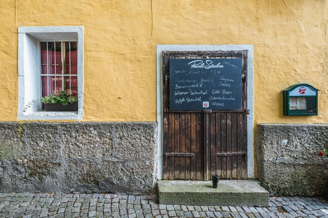 A chalkboard menu for Pauli Stubm hangs above the weathered wooden door of the tavern's ochre-yellow wall in Salzburg, next to a barred window with red curtains and a geranium box.