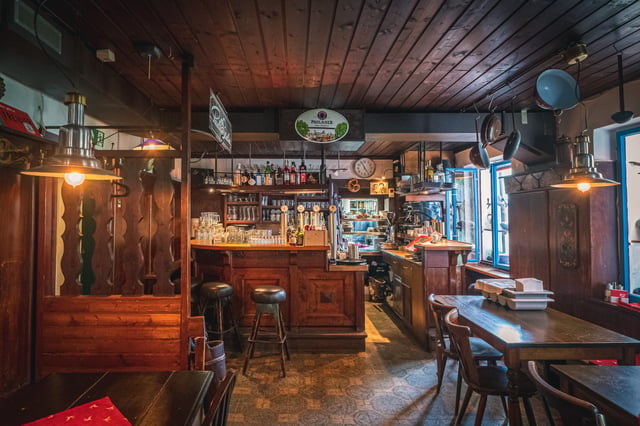 The wood-panelled interior of Pauli Stubm looks across bar stools to the counter, with taps, a Paulaner sign, hanging pans, enamel lamps and a blue-framed window to the right.