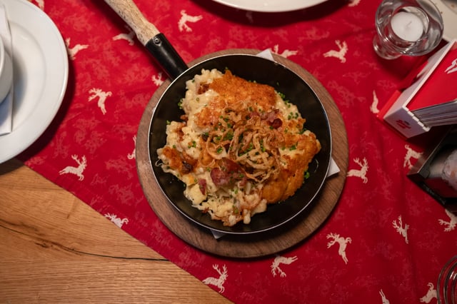 A black pan of Kasnocken cheese spaetzle topped with crisp fried onions, bacon and chives, set on a wooden board and photographed from directly above on a red deer-patterned runner, with a tealight and matchbox alongside.