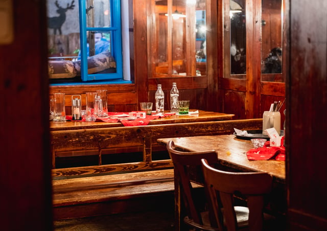 The wood-panelled Stuberl room at Pauli Stubm, with a bright blue-framed interior window, a deer silhouette on the wall beyond, long benches, empty glasses and red deer-patterned runners on the scrubbed tables.