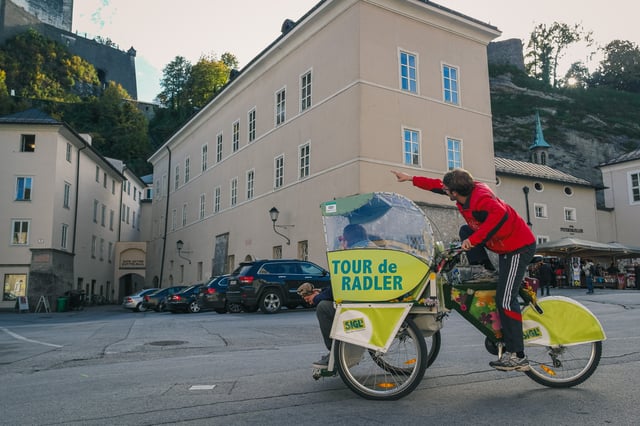 A Rikscha Tours driver in a red jacket stands on the pedals of his green Tour de Radler rickshaw and points ahead, on Kapitelplatz below the walls of Hohensalzburg fortress.